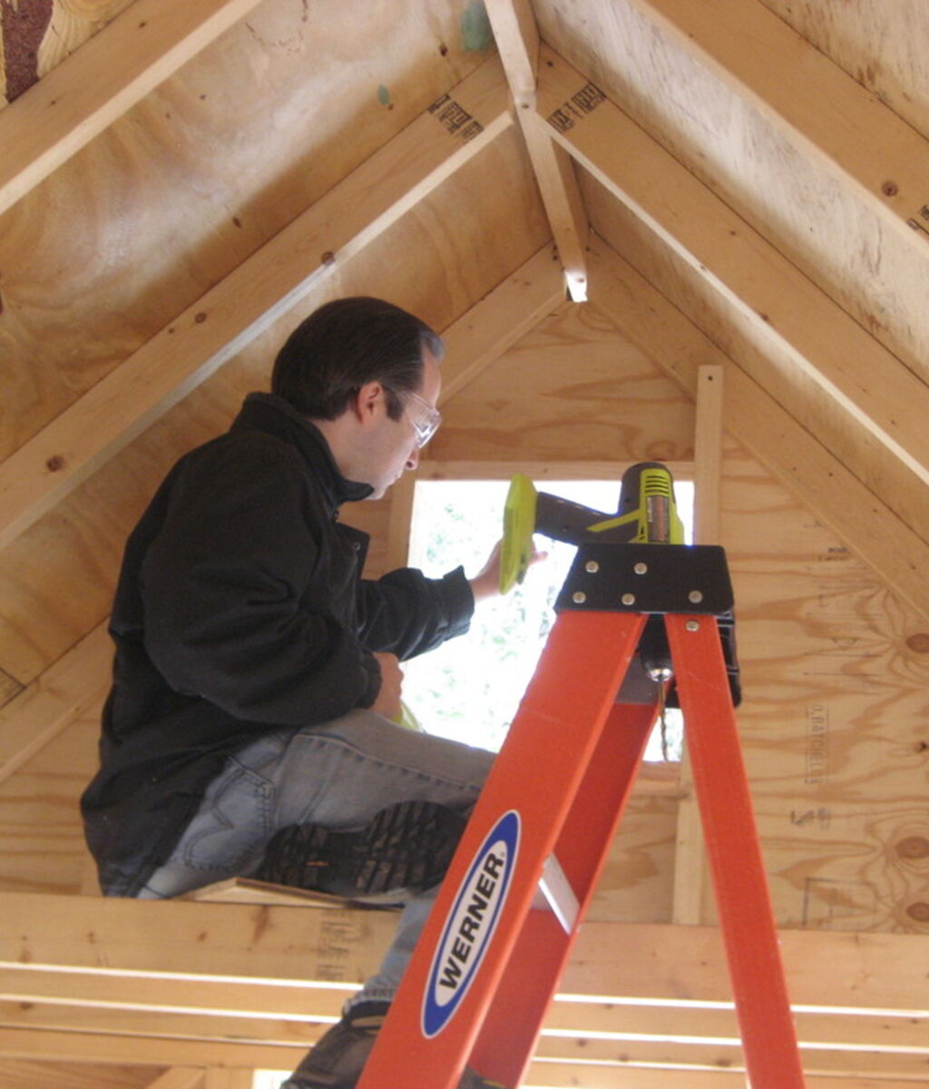 A carpenter on an orange stepladder working on the framing of a bright timber attic