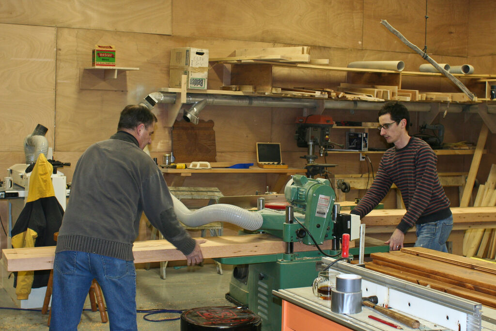 Two craftsmen working at machines in a busy woodworking shop full of timber and tools